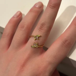 Ring från &other stories - Elegant guldfärgad ring med två öppna ändar dekorerade med små gröna stenar. Justerbar storlek. 