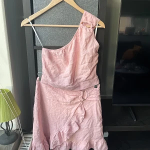 Rosa oneshoulder klänning - Säljer en söt rosa oneshoulder klänning med volangdetaljer. Klänningen har en unik design med en ringdetalj vid axeln och midjan. Perfekt för sommarens festligheter!