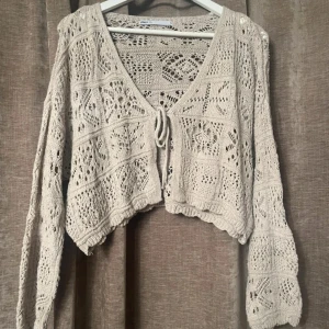 Beige stickad kofta med knyt - Säljer en beige stickad kofta med v-ringning och knyt framtill. Koftan har ett vackert hålmönster och långa ärmar. Perfekt för lager-på-lager-stil.