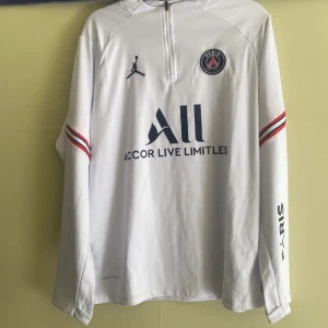  långärmad fotbollströja från Jordan x PSG - Snygg vit långärmad fotbollströja från Jordan x PSG med klubbens emblem på bröstet. Tröjan har röda och svarta detaljer på ärmarna och en halv dragkedja framtill. Perfekt för fotbollsfans som vill ha stil och komfort.