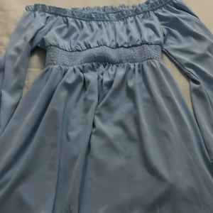 Blå offshoulder klänning - Charmig blå offshoulder klänning med volangdetaljer. Klänningen har en smockad midja som ger en fin passform och långa ärmar. Perfekt för sommarens alla tillfällen.