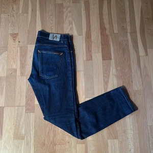 Mörkblå jeans från Nudie Jeans - Snygga mörkblå jeans från Nudie Jeans med klassisk femficksdesign och orange sömmar. Jeansen har en slim passform. Modellen heter grim Tim 