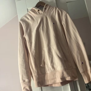 Beige hoodie från Champion - Säljer en beige hoodie från Champion med klassisk logga på bröstet och ärmen. Tröjan har en känguruficka och är tillverkad i mjukt material för extra komfort. Perfekt för en avslappnad stil. (Den är mer rosa än vad den ser ut på bild)