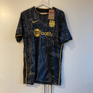 Svart och gul FC Barcelona fotbollströja från Nike - Snygg FC Barcelona tröja i svart med gula detaljer och ett unikt mönster. Tröjan har korta ärmar och klubbens emblem samt sponsorlogga på bröstet. Perfekt för fans som vill visa sitt stöd med stil. Special edition 23/24