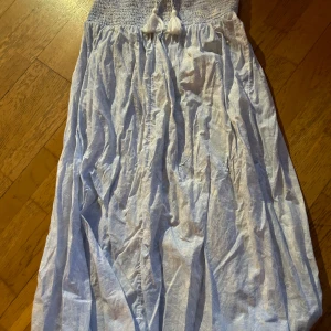 Ljusblå långklänning med tofsar - Charmig ljusblå långkjol med smockdetaljer och dekorativa tofsar framtill. Klänningen har ett subtilt mönster och är perfekt för varma dagar. VIKTIGT( Den finns en liten curry fläck på framsidan som däremot går att ta bort)