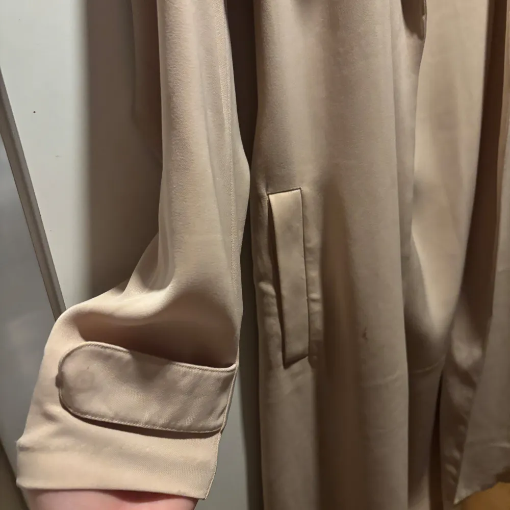 Snygg beige trenchcoat med knytband i midjan och klassisk krage. Har en liten fläck. Takit.