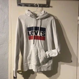Vit hoodie från Levi's - Säljer en klassisk vit hoodie från Levi's med deras ikoniska logga i blått och rött på bröstet.