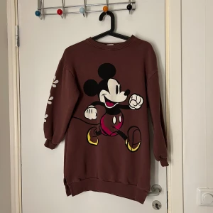 Brun tröja med Musse Pigg-motiv - Säljer en brun tröja med ett stort Musse Pigg-tryck på framsidan. Tröjan har långa ärmar med vita blommor längs ena ärmen. Perfekt för en avslappnad stil.