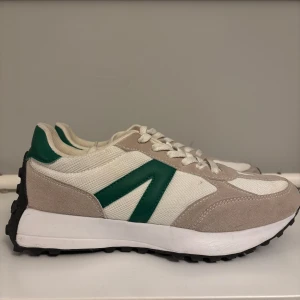 Beige och gröna sneakers - Snygga sneakers i beige och grönt med vit sula och svart mönstrad undersida. Skorna har snörning och en stilren design med gröna detaljer på hälen och sidan.
