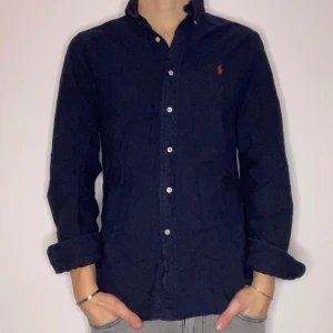 Ralph lauren skjorta - Snygg mörkblå skjorta från Ralph Lauren i slim fit. Skjortan har långa ärmar och knappar framtill. Den är broderad med det klassiska röda logotypen på bröstet. Perfekt för en stilren look. Modellen är 180 cm lång och bär storlek S. Vid övriga funderingar eller frågor är det bara att höra av sig!🙌