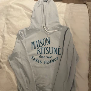 Ljusblå hoodie från Maison Kitsuné - Säljer en stilren ljusblå hoodie från Maison Kitsuné med tryck 'Palais Royal Paris, France' i mörkblått. Tröjan har en klassisk passform. Perfekt för en avslappnad look. Enbart använd 2 gånger, säljer eftersom den är för liten! Nypris ca 3000
