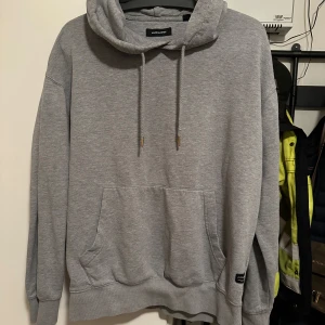 Grå hoodie från Jack & Jones - Säljer en stilren grå hoodie från Jack & Jones. Den har en klassisk känguruficka och justerbar dragsko i huvan. Perfekt för en avslappnad look. Noppor eller ytludd kan förekomma.