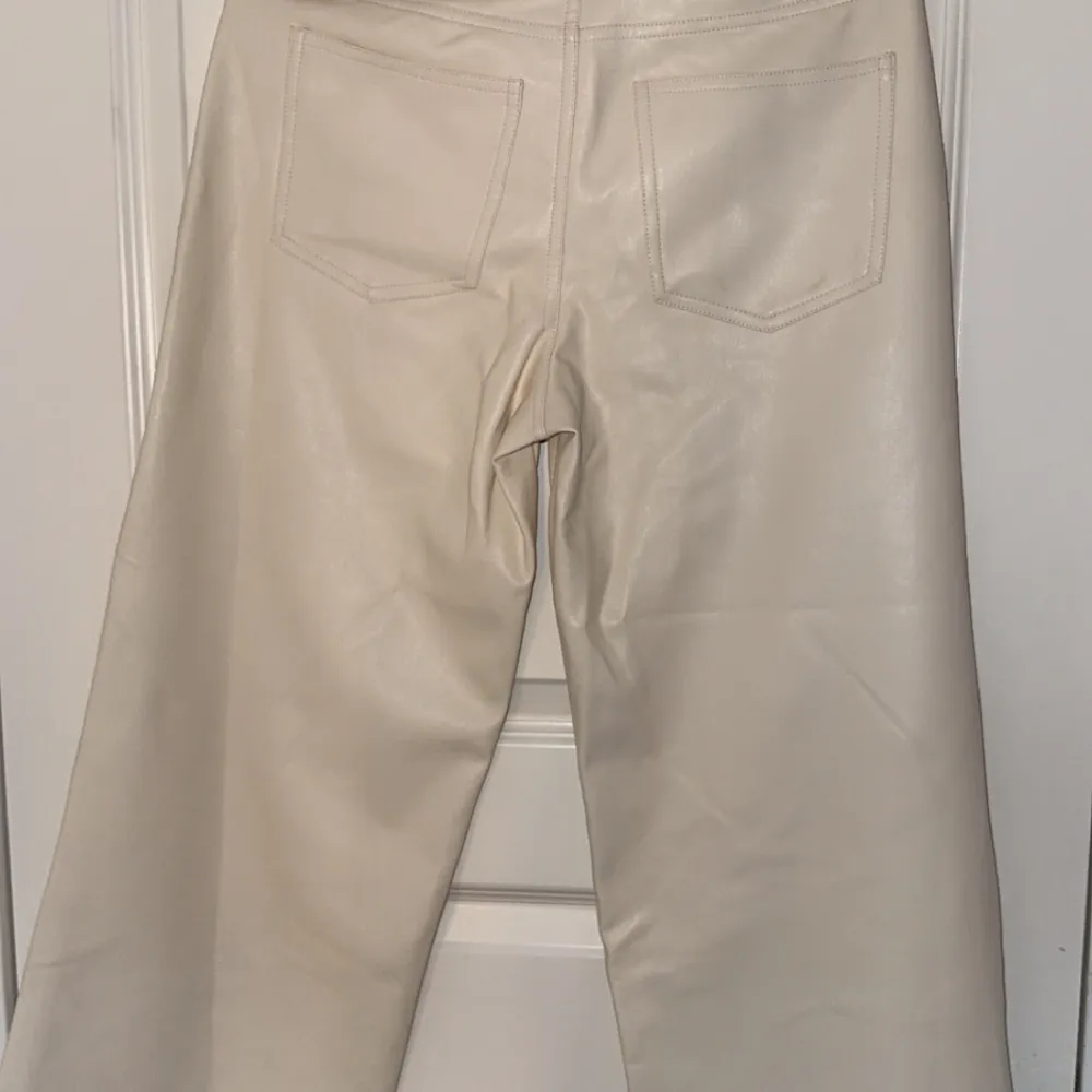 Snygga beige byxor i syntetmaterial med hög midja och raka ben. Perfekta för en stilren look. Byxorna har fickor bak och en klassisk design framtill.. Farkut & Housut.