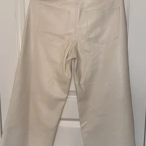 Beige byxor i syntetmaterial - Snygga beige byxor i syntetmaterial med hög midja och raka ben. Perfekta för en stilren look. Byxorna har fickor bak och en klassisk design framtill.