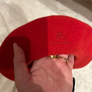 Röd basker  Burberry - Snygg röd basker ( vet inte om den är äkta!!) , tillverkad i 100% ull. Perfekt för att ge en touch av stil till din outfit. Tillverkad i England.