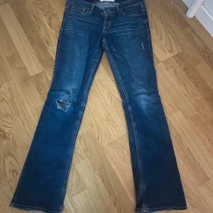 Blå bootcut lowaist jeans från Hollister - Säljer ett par blå bootcut jeans från Hollister med slitningar på benen. De har en klassisk femficksdesign och knappgylf. Perfekta för en avslappnad stil.