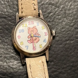 Barnklocka med nallemotiv från Louis Picard - Charmig barnklocka med ett sött nallemotiv på urtavlan och färgglada siffror. Klockan har ett beige armband med krokodilmönster och boett i rostfritt stål. Perfekt för barn som älskar djur och färger!