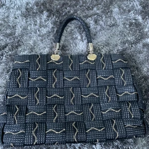 Svart handväska med mönster - Elegant svart handväska med ett unikt vävt mönster och guldiga detaljer. Väskan har ett flätat handtag och flera fack med dragkedja för praktisk förvaring. Perfekt för att ge en stilfull touch till din outfit.
