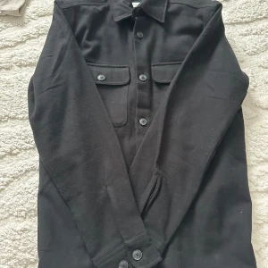 Svart långärmad skjorta/jacka - Snygg svart långärmad skjorta med knappar och två bröstfickor. Perfekt för en stilren look. Skjortan har en klassisk krage och är tillverkad i ett mjukt material.