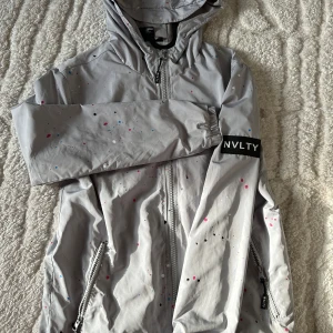 Grå vindjacka med färgstänk från NVLTY - Snygg grå vindjacka från NVLTY med färgglada stänk i rosa och blått. Jackan har en praktisk huva och dragkedja framtill. Perfekt för blåsiga dagar.