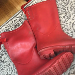 Röda stövlar med snörning - Snygga röda stövlar med dekorativ snörning och rosett. Perfekta för regniga dagar och ger en färgklick till din outfit.