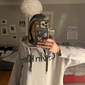 Grå hoodie från Calvin Klein - Säljer en snygg grå hoodie från Calvin Klein med svart logga på framsidan. Tröjan har en justerbar huva med svarta snören och långa ärmar. Perfekt för en avslappnad stil.