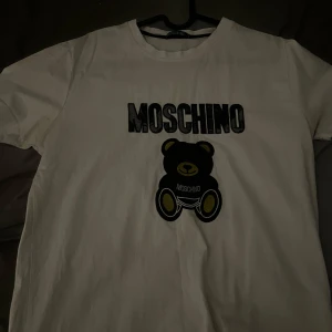  Moschino  - Snygg vit t-shirt från Moschino med ett stort svart och gult nalletryck på framsidan. Perfekt för en casual look med en touch av lyx. T-shirten har en rund halsringning och korta ärmar. Vet inte om de är 1 :1 köpt av en polare men den är Rikigt skön, kom me prisförslag 