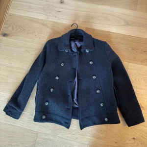 Skepparjacka från GAP - Säljer en stilren mörkgrå kappa från GAP i ull. Kappan har dubbelknäppning med stora knappar och en klassisk krage. 🤍