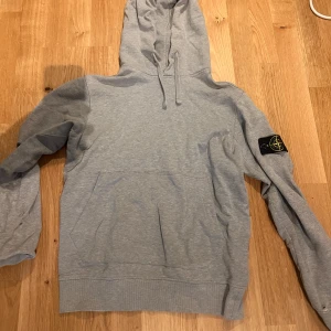 Grå hoodie från Stone Island - Säljer en stilren grå hoodie från Stone Island med klassisk logga på ärmen. Tröjan har en bekväm passform med känguruficka och justerbar huva. Perfekt för en avslappnad look. Den är äkta och går att autentiseras. Den har dock ett hål vid armbågen, annars är den i bra skick.