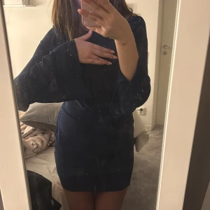 Mörkblå glittrig klänning - Snygg mörkblå klänning med glittrande detaljer och öppen rygg. Klänningen har långa ärmar och en kort längd, perfekt för en festlig kväll. Den har en elegant knytning i ryggen som ger en extra touch. Den populära parthea klänningen. Använd en gång