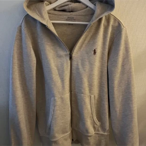 Grå hoodie från Ralph Lauren - Säljer en stilren grå hoodie från Ralph Lauren med dragkedja och klassisk logga på bröstet. Perfekt för en avslappnad look. Den har en bekväm passform och praktiska fickor framtill.
