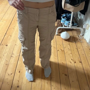 Low waisted beige cargobyxor - Snygga lågmidjade beige cargobyxor från H&M Divided med flera praktiska fickor. Byxorna har en loose passform och är perfekta för en avslappnad stil. De är tillverkade i ett bekvämt material och har en elastisk midja för extra komfort. Storlek: 34