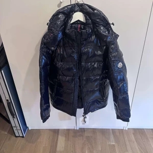 Moncler jacka - Snygg svart pufferjacka från Moncler med glansig yta och dragkedja. Jackan har en hög krage och långa ärmar, perfekt för kyliga dagar. Den har också en ikonisk Moncler-logga på ärmen. Väldigt populär jacka.