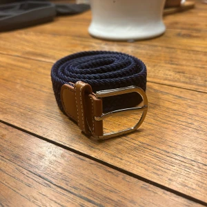 Blått flätat bälte med bruna detaljer - Snyggt blått flätat bälte med bruna läderdetaljer och en stilren silverfärgad spänne. Perfekt för att ge en casual look en extra touch. Passar till både jeans och chinos.