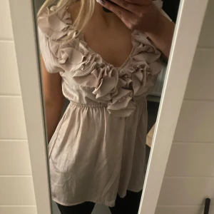 Beige blus med volangdetaljer - Säljer en söt beige blus med volangdetaljer runt den v-ringade halsen. Blusen har korta ärmar och en elastisk midja som ger en fin passform. Perfekt för en stilren och romantisk look.😍