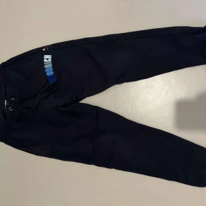 Svarta mjukisbyxor från Hoodrich - Snygga svarta mjukisbyxor från Hoodrich med broderad logga i blått och vitt. Byxorna har snörning i midjan och en bekväm passform. Perfekta för en avslappnad stil.