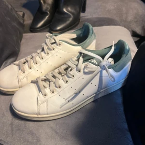 Vita Adidas Stan Smith sneakers - Klassiska vita Adidas Stan Smith sneakers med gröna detaljer och snörning. Perfekta för en stilren och tidlös look.