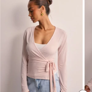 Ljusrosa omlottopp - Säljer en stilren ljusrosa omlottopp med knytband i midjan. Toppen har långa ärmar och en v-ringad design som ger en elegant look. Perfekt för att matcha med jeans eller kjol. Slutsåld på hemsidan.