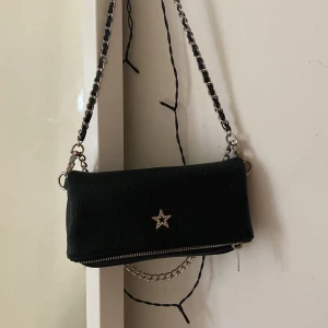 Svart axelväska från Gina Tricot - Snygg svart axelväska från Gina Tricot med en stjärndetalj i silver på framsidan. Väskan har en kedjerem och dragkedja för säker förvaring. Perfekt för att ge din outfit en cool touch.