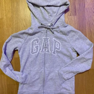 Grå hoodie från GAP - Säljer en klassisk grå hoodie från GAP med dragkedja och stor logga på framsidan. Tröjan har en bekväm huva med dragsko och långa ärmar. Perfekt för en avslappnad stil. 🩶