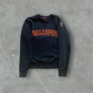Svart sweatshirt från Parajumpers - Säljer en svart sweatshirt från Parajumpers med orange text på framsidan. Tröjan har långa ärmar och en rund halsringning. Perfekt för en avslappnad stil.  
