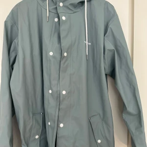 Ljusblå regnjacka från Tretorn - Säljer en stilren grå regnjacka från Tretorn i storlek large. Jackan har en praktisk huva med dragsko och stängs med både dragkedja och knappar. Perfekt för regniga dagar! Liten fläck se bild