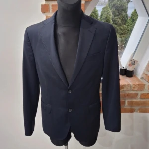 Kavaj från Oscar Jacobson - Elegant blå kavaj från Oscar Jacobson med klassisk design. Kavajen har två knappar framtill och är tillverkad i ett stilrent material. Perfekt för formella tillfällen. Gjord i ett tunnare material så passar utmärkt för sommaren. Mycket fint skick. Storlek 48 motsvarar S/M 