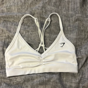 Vit sporttopp från Gymshark - Säljer en vit sporttopp från Gymshark med smala axelband och snygg snörning i ryggen. Toppen har en v-ringad design och är perfekt för träning. Materialet är mjukt och stretchigt för bästa komfort.