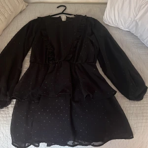 Svart klänning från Gina Tricot - Snygg svart klänning från Gina Tricot med långa ärmar och volangdetaljer. Klänningen har ett diskret glittrigt mönster som ger en extra touch. Perfekt för en stilren look.