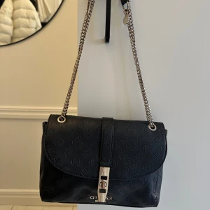Svart axelväska från Guess - Snygg svart axelväska från Guess med kedjeaxelrem och präglat mönster. Väskan har en elegant låsdetalj framtill och Guess-logotypen i silver. Perfekt för att ge din outfit en stilfull touch.  Mått: bredd 25cm höjd 18cm