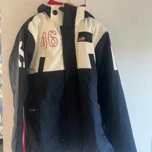 Svart och vit jacka från Sebago - Säljer en cool svart och vit jacka från Sebago med röda detaljer och nummer 46 på bröstet. Jackan har en stor huva och dragkedja framtill. Perfekt för kyliga dagar med sin stilrena design och amerikanska flagga på bröstet.
