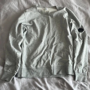 Grå sweatshirt från C.P. Company - Säljer en stilren grå sweatshirt från C.P. Company. Tröjan har en rund halsringning och en cool detalj med en svart lins på ärmen. Perfekt för en avslappnad look.