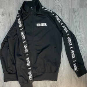 Svart Zip-Hoodie Hoodrich - Snygg svart Zip-Hoodie från Hoodrich med dragkedja och logga längs ärmarna. Tröjan har en hög krage och ribbade muddar.