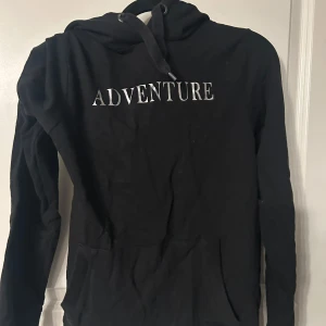 Svart hoodie med texten 'ADVENTURE' - Säljer en svart hoodie med texten 'ADVENTURE' i vitt på framsidan. Tröjan har en klassisk design med huva och långa ärmar. Perfekt för en avslappnad stil.